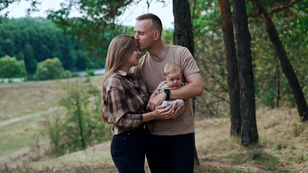 Happy young parents with their firstborn baby. Caucasian couple stand in nature with their newborn. Man kisses his wife on the forehead.