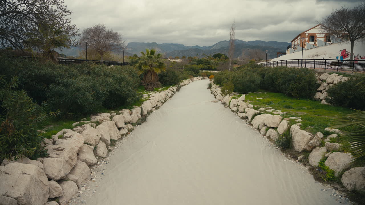 Vista nuvolosa di acqua piovana torbida che scorre attraverso un canale di drenaggio urbano rivestito in pietra, circondato da vegetazione ed edifici residenziali. Deflusso di acque piovane in Spagna