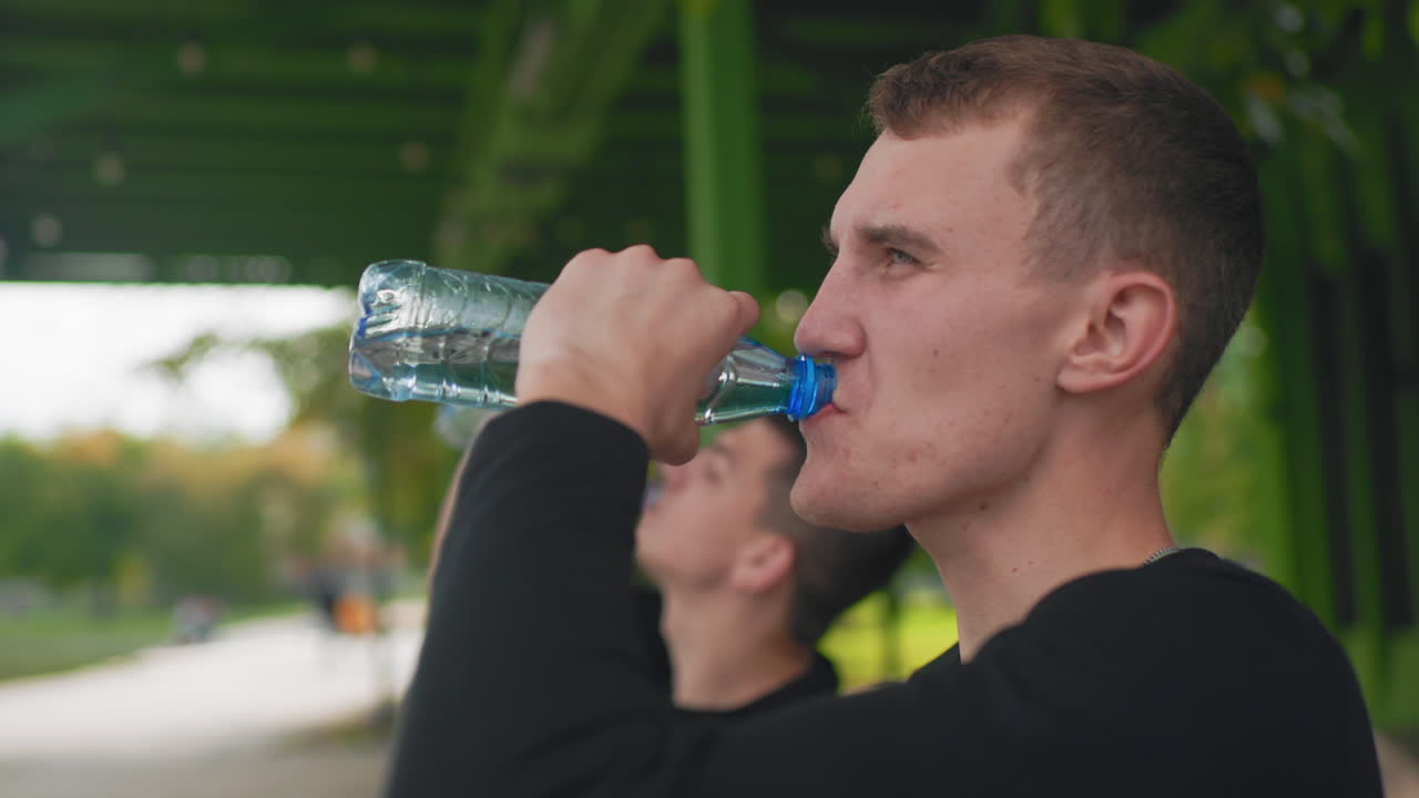 Белый мужчина-спортсмен пьет воду на открытом воздухе, отдыхая под зеленым павильоном после тренировки, друг на размытом фоне, крупный план глотка из бутылки и пота, повседневная черная футболка, выражение облегчения