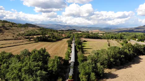 aerial: old diesel train traveling along fields under summer blue sky with some clouds on a sunny day