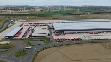 Aerial drone orbiting footage of large industrial warehouse distribution centre, with heavy goods trucks loading and unloading, wind turbine power plant renewable energy, Rugby England UK