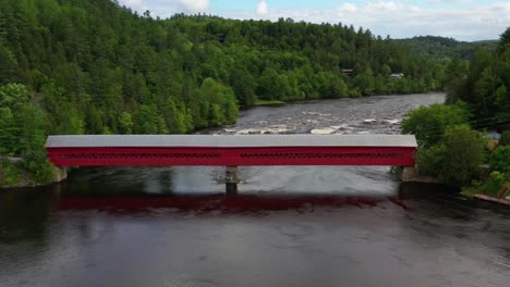 Tiro-Aéreo-Desde-Una-Plataforma-Rodante-Hacia-Un-Puente-De-Madera-Cubierto-De-Color-Rojo-Brillante-Sobre-Un-Río-Canadiense-Con-Rápidos-Río-Abajo