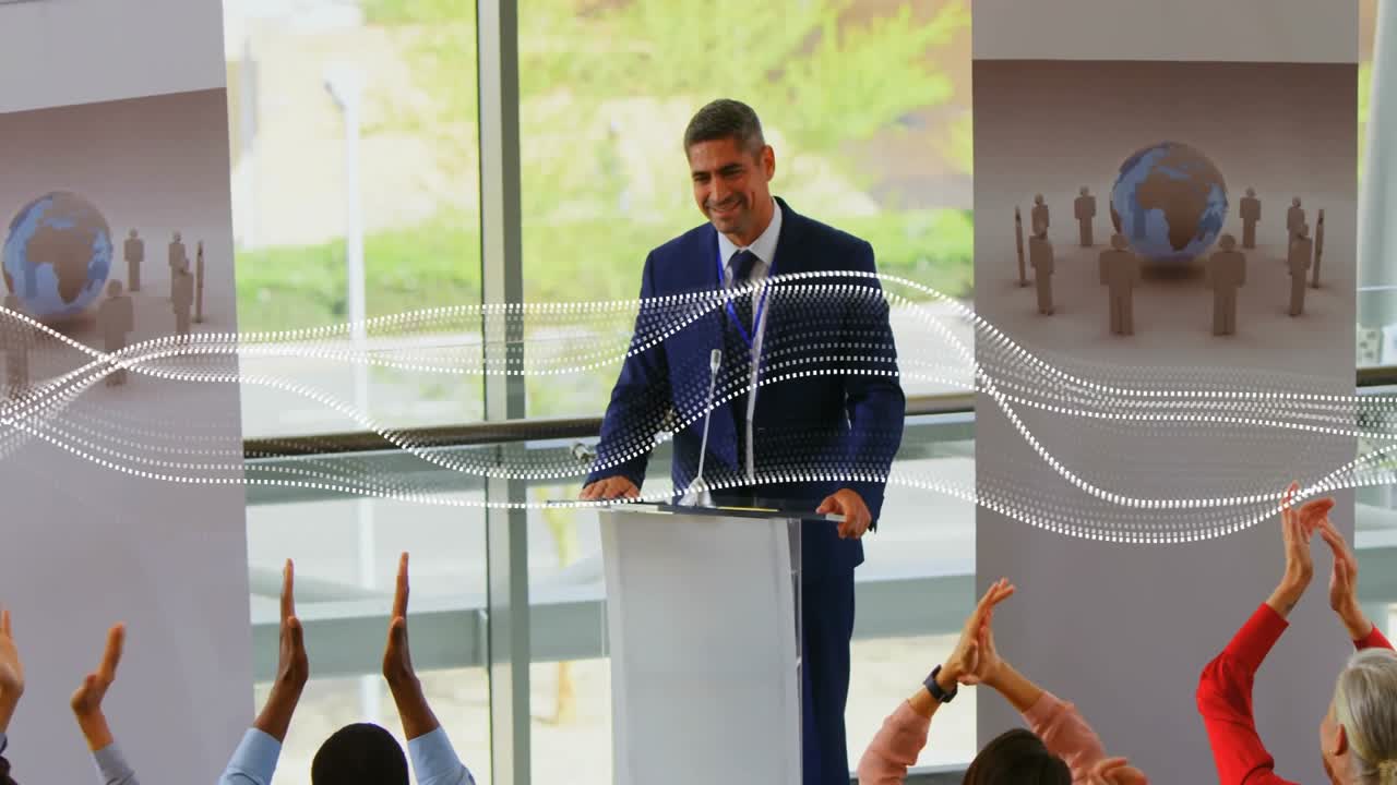 Apresentador masculino cumprimentando a plateia no pódio em evento de negócios, recebendo aplausos, assentindo com polegar para cima no final