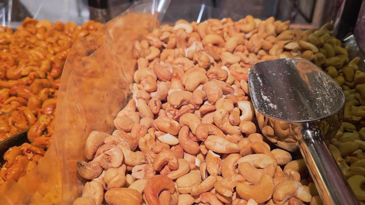 Utstilling av ristede cashewnøtter og blandede nøtter i et spesialmatmarked, med serveringsskjeer i rustfritt stål, klare plastbrett, saltet tekstur og varm butikkbelysning for å fremheve snacks.