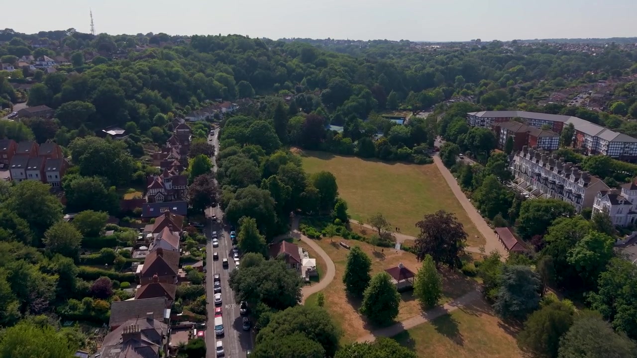 Penerbangan drone maju melintasi Taman Alexandra di Hastings, dengan rumputnya yang kering kecoklatan selama gelombang panas musim panas, dengan jalan-jalan bertingkat, lalu lintas yang mengantre, dan apartemen modern yang berbatasan dengan taman yang dipenuhi pepohonan