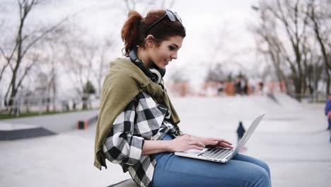 una joven morena de moda está sentada en un banco con una computadora portátil de rodillas, escribiendo. auriculares en el cuello, gafas. en el parque los niños patinan en scooters y bicicletas. vista lateral