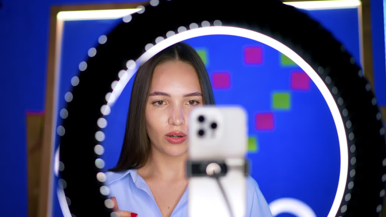 Smiling Caucasian woman stands behind the phone on tripod and light ring. Positive blogger taking video.
