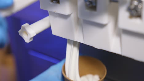 hand in blue gloves puts a paper cup under a food automat. soft creamy ice-cream is filling the cup close up.