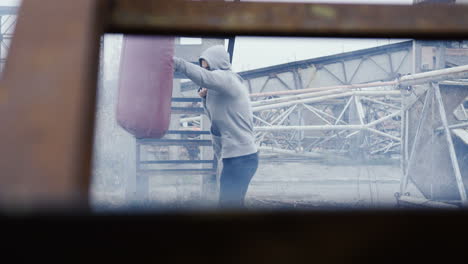 caucasian man in grey hoodie hitting a punching bag outdoors an abandoned factory on a cloudy morning