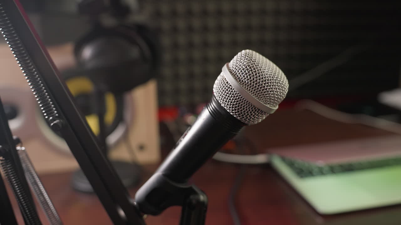 fotografía de primer plano de un micrófono en un soporte con un fondo borroso con un altavoz, auriculares y una computadora. la escena captura los elementos esenciales de un estudio de grabación