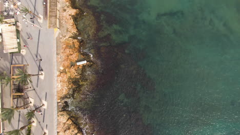 Tropical-city-coastline-with-people-walking-and-majestic-ocean-water,-aerial-top-down-view