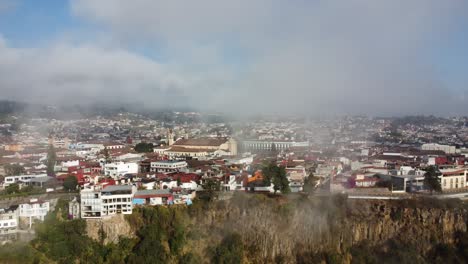 Drone-shot-of-historic-center-of-Zacatlan-filled-with-traditional-houses-red-tile-roofs,-church-buildings,-and-public-gardens-through-clouds,-Atmospheric-cityscape-in-clouds,-Puebla,-Mexico