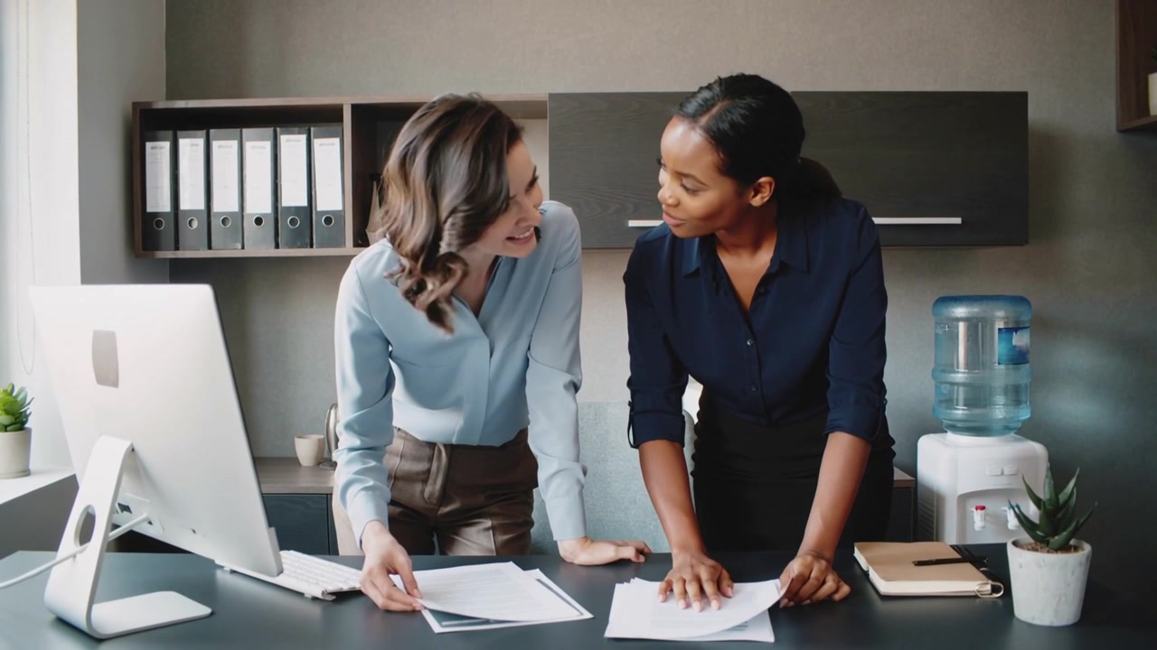 Sorting two coworkers in blouses leaning at desk after docs placed arranging papers for meeting