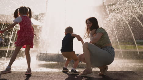 Little-girl-jumps-mother-holds-boy-touching-fountain-water