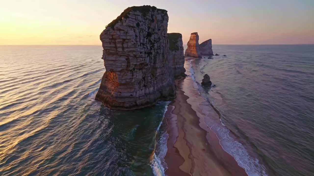 Video aéreo que captura acantilados dramáticos al atardecer, mostrando serenas olas oceánicas y una playa de arena