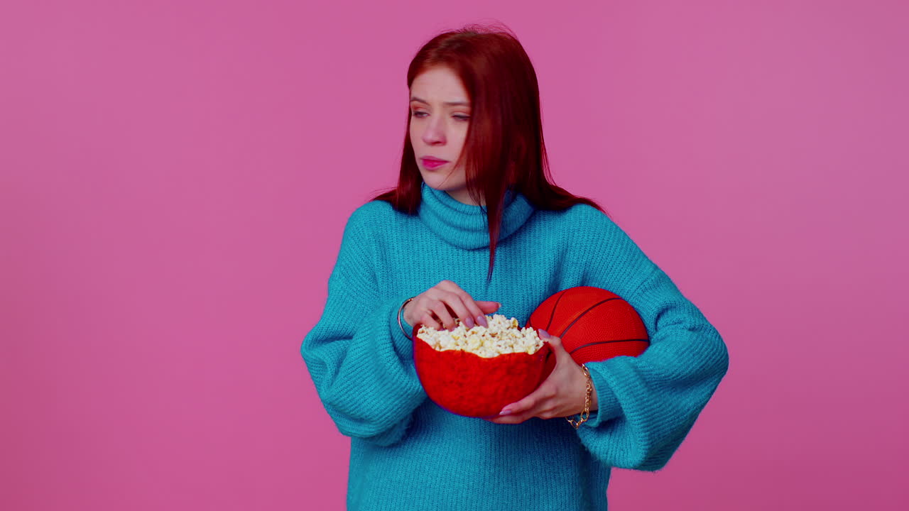 красноволосая молодая баскетбольная фанатка ест попкорн, делает жест победителя, празднует победу.