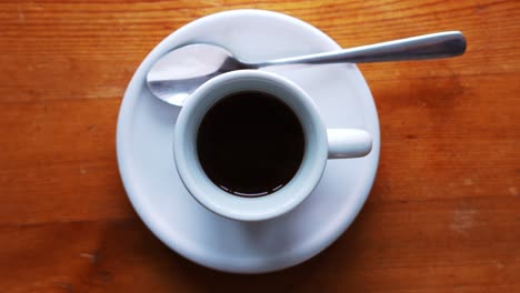 zoom down shot on a hot espress cup standing on a white plate with spoon and brown wooden table