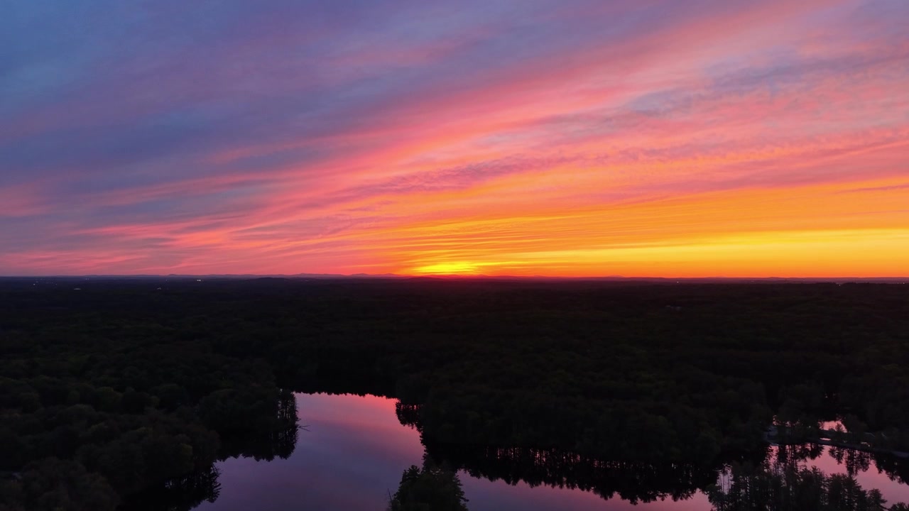 Dronebillede af farverig lyserød og orange solnedgangshimmel, der spejler sig i en rolig sø omgivet af mørk skov i Harold Parker State Forest, Massachusetts. Plads til tekst