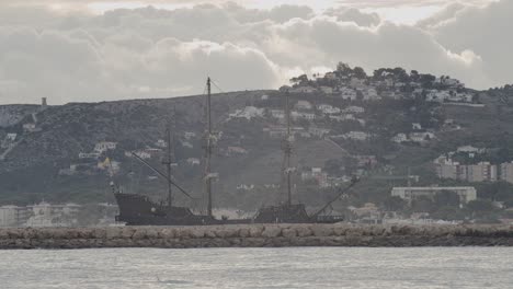 Galeone-Aus-Dem-16.-Jahrhundert,-Andalusisches-Nachbauschiff,-Das-An-Einem-Schönen-Bewölkten-Tag-Bei-Sonnenaufgang-Hinter-Einem-Wellenbrecher-Mit-Berg-Und-Häusern-Im-Hintergrund-Im-Hafen-Ankommt