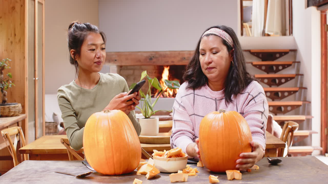 Multiracial grandmother and young woman carving pumpkins, capturing memories at home, halloween time
