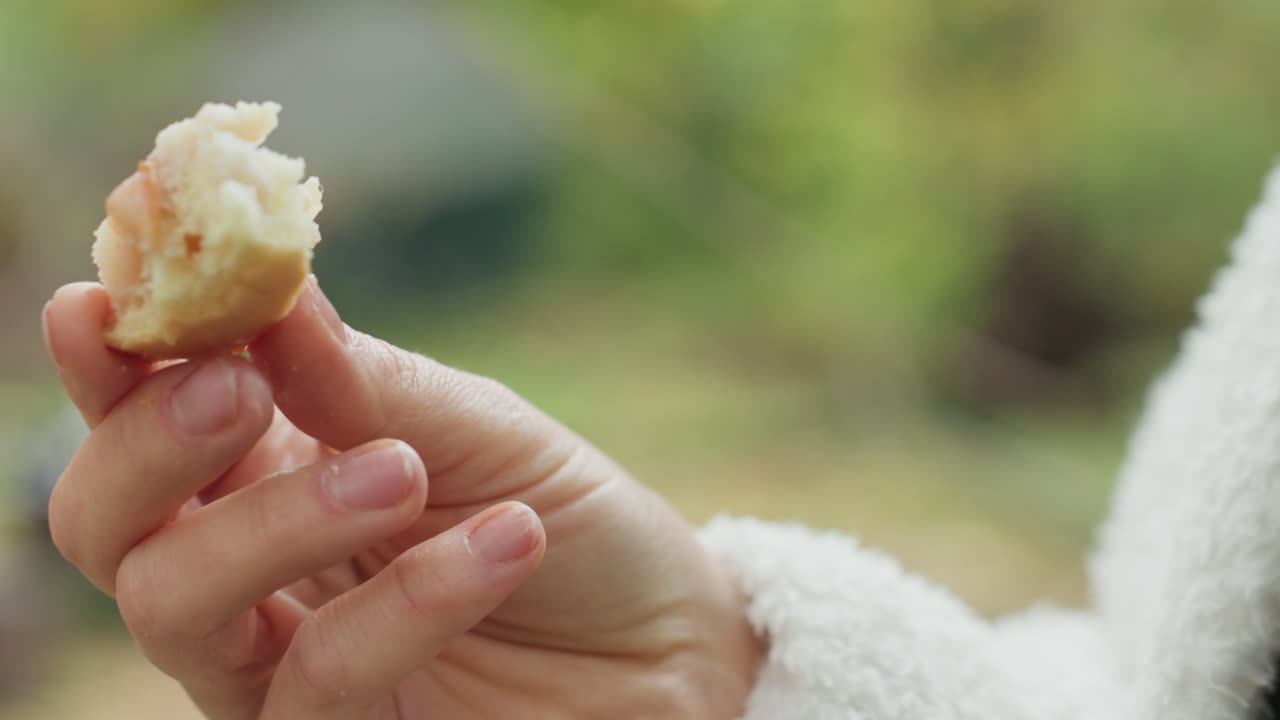 Donna caucasica che assaggia il pane all'aperto in un giardino, primo piano sulla mano e sulle labbra, mollica rustica e consistenza burrosa, atmosfera rilassata, dettaglio accogliente dell'accappatoio, luce mattutina e atmosfera serena