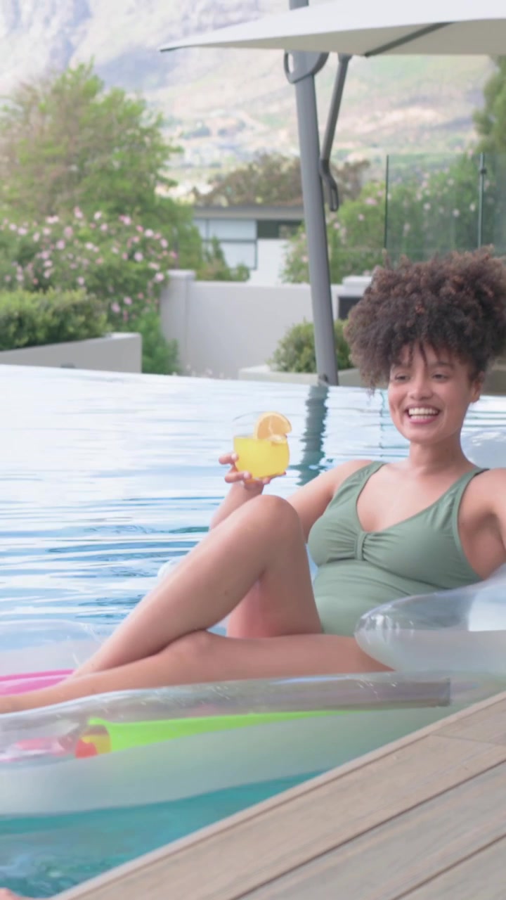 Vertikales Video: Frau in grünem Badeanzug auf Pool-Schwimmhilfe, hebt Getränk und lächelt, als eine Hand erscheint.