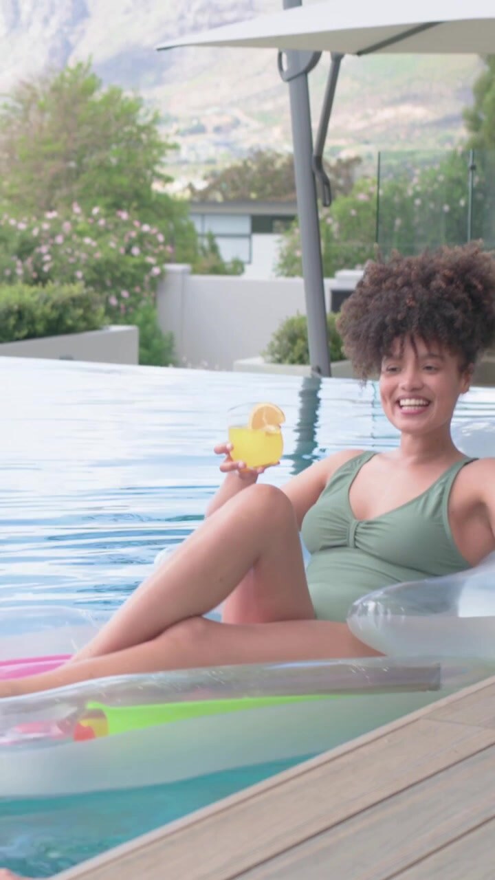 Vertical video: Woman in green swimsuit on pool float, lifting drink and smiling at hand appearing