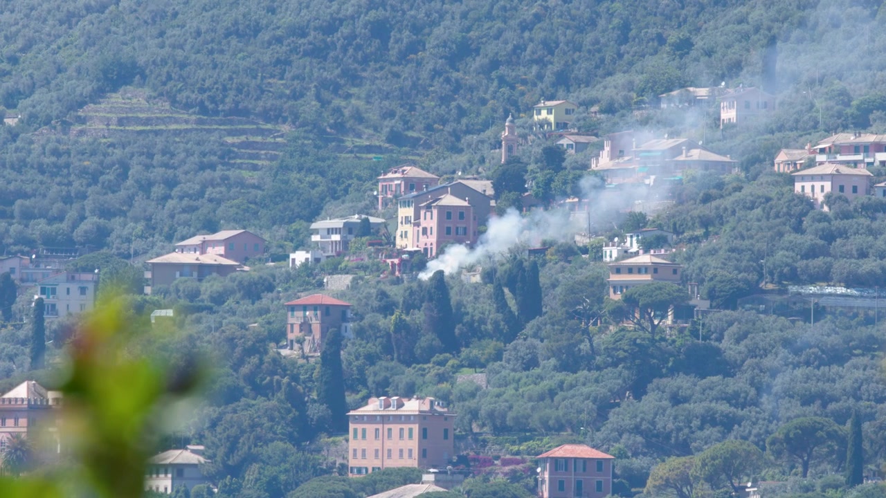 Inquadratura ampia statica di fumo che si disperde da case colorate sulla collina annidate tra rigogliosi ulivi