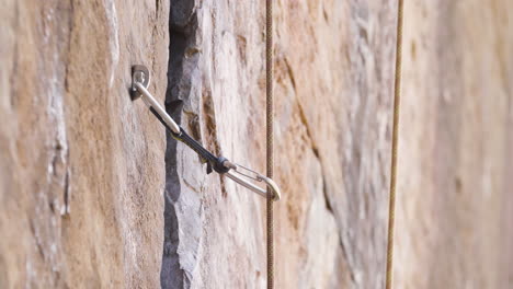 Close-up-shot-of-carabiner-secured-to-a-piton-against-rock-face-with-rock-climbing-rope-feeding-through