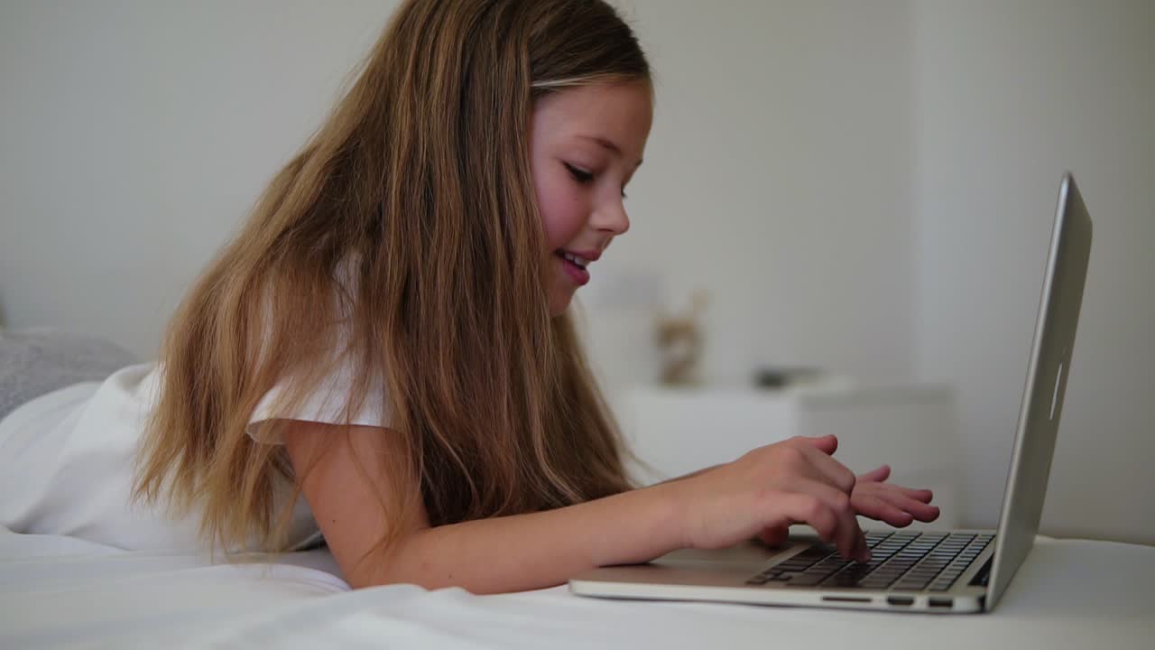 primer plano de una hermosa chica europea de cabello largo escribiendo en una computadora portátil. una niña pequeña sonriente con interés mira la computadora portátil y se acuesta en la cama. vista lateral, cámara lenta