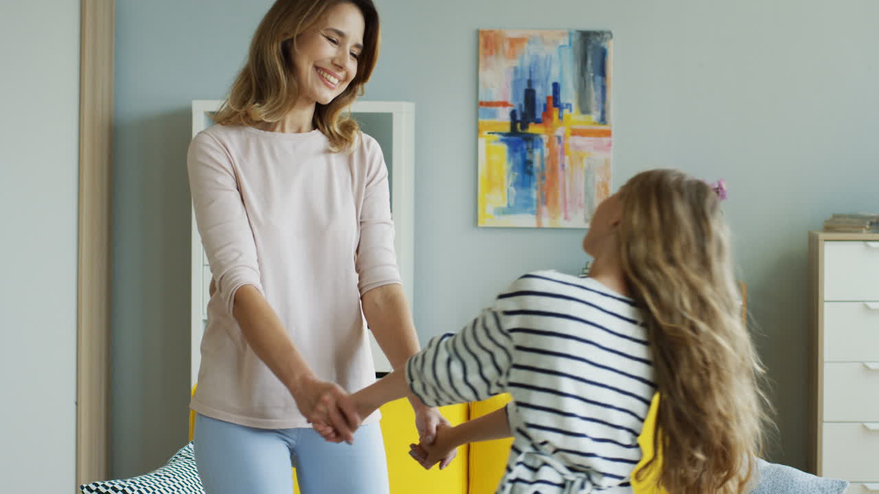 feliz madre relajada e hija pequeña tomándose de la mano y bailando en la sala de estar