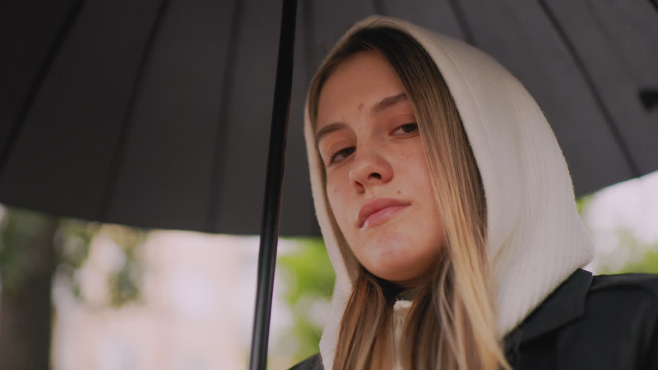 Close-up of woman in hooded coat holding umbrella outdoors on rainy autumn day, showing detail of hands, clothing, and blurred natural background creating moody seasonal atmosphere