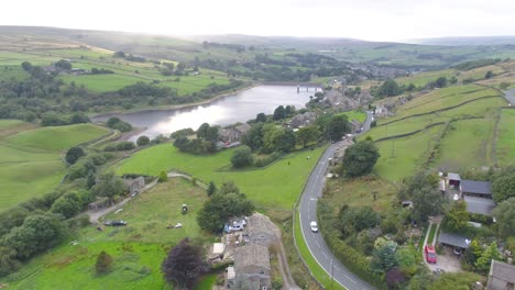 Drone-footage-panning-from-the-bottom-of-a-small-Yorkshire-countryside-valley-to-the-top-of-the-hill,-including-village-stone-houses,-farms,-country-roads,-dry-stone-walls,-reservoir-and-moors