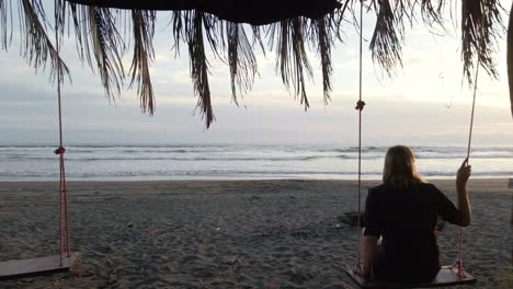 drone flight to the ocean through a palm leaf covered pergola with inside a lady on a swing on the beach of playa bandera in costa rica