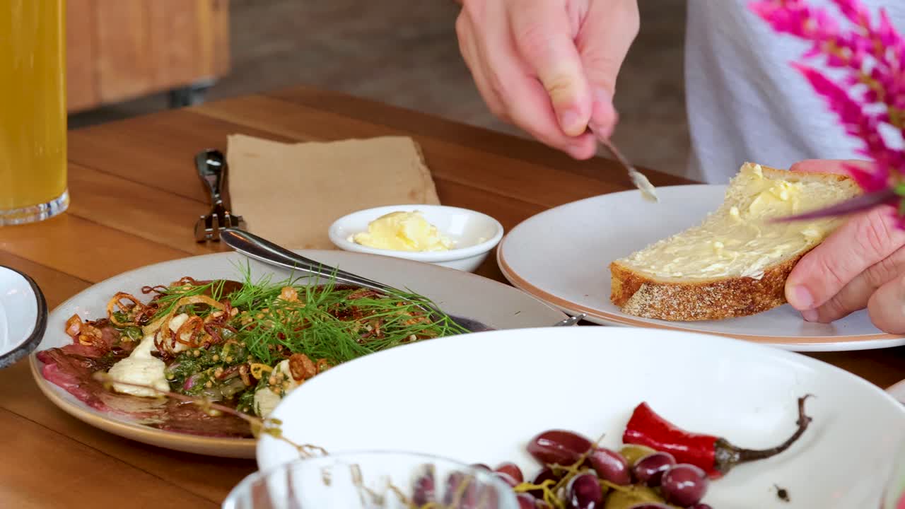 A person spreads butter onto a slice of bread at a well-lit breakfast table with fresh fruit, herbs, and juice. Close-up, steady camera