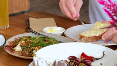 a person spreads butter onto a slice of bread at a well-lit breakfast table with fresh fruit, herbs, and juice. close-up, steady camera