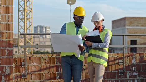 arquitecta caucásica con documentos en la carpeta en las manos hablando y discutiendo el trabajo con un hombre afroamericano constructor que le muestra algunos borradores y planes.