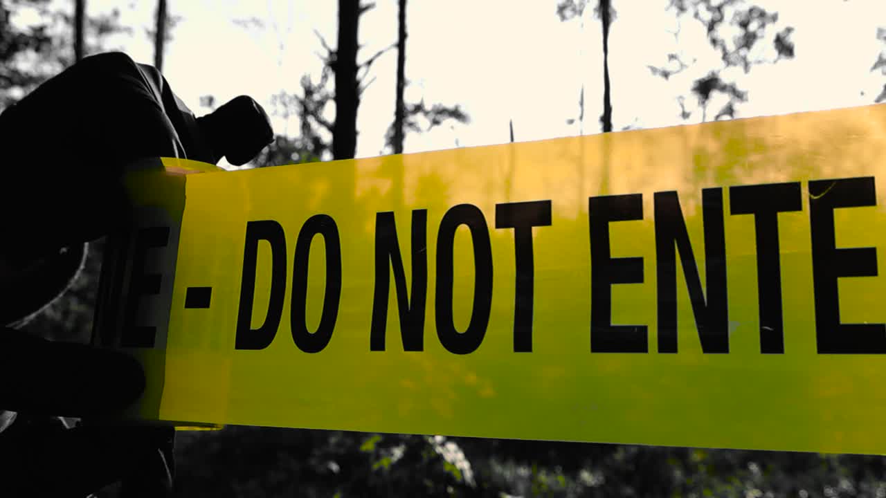 Policeman investigator with black nylon protective gloves unrolling or unraveling a yellow colored crime scene police caution tape in front of a dark forest woodland area while sun is lighting it