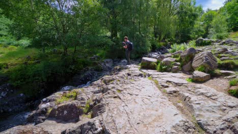 Kleiner-Junge-Fotografiert-Einen-Langsam-Fließenden-Waldbach-Mit-Farnen-Am-Flussufer-Und-Wasser,-Das-über-Die-Felsen-Fließt