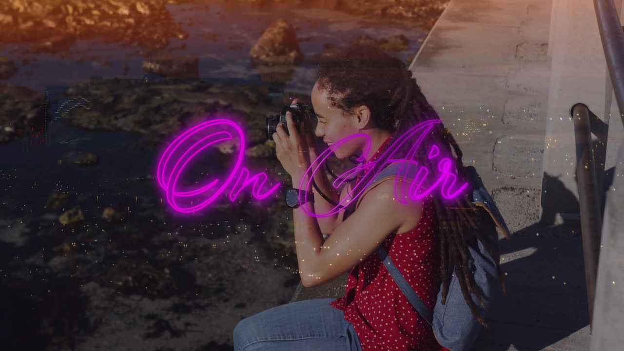 Photographer broadcasting live media by rocky shoreline, showing camera, neon On Air overlay