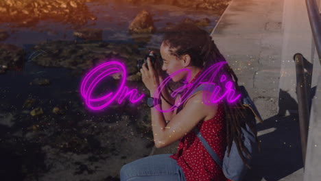photographer broadcasting live media by rocky shoreline, showing camera, neon on air overlay
