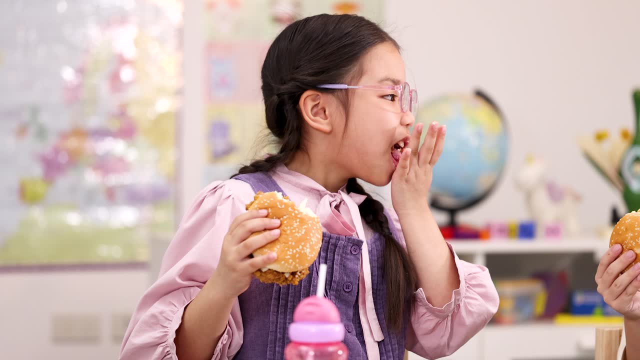 Elementary schoolgirl enjoys chicken burger at classroom lunch table, bright lighting, static medium shot