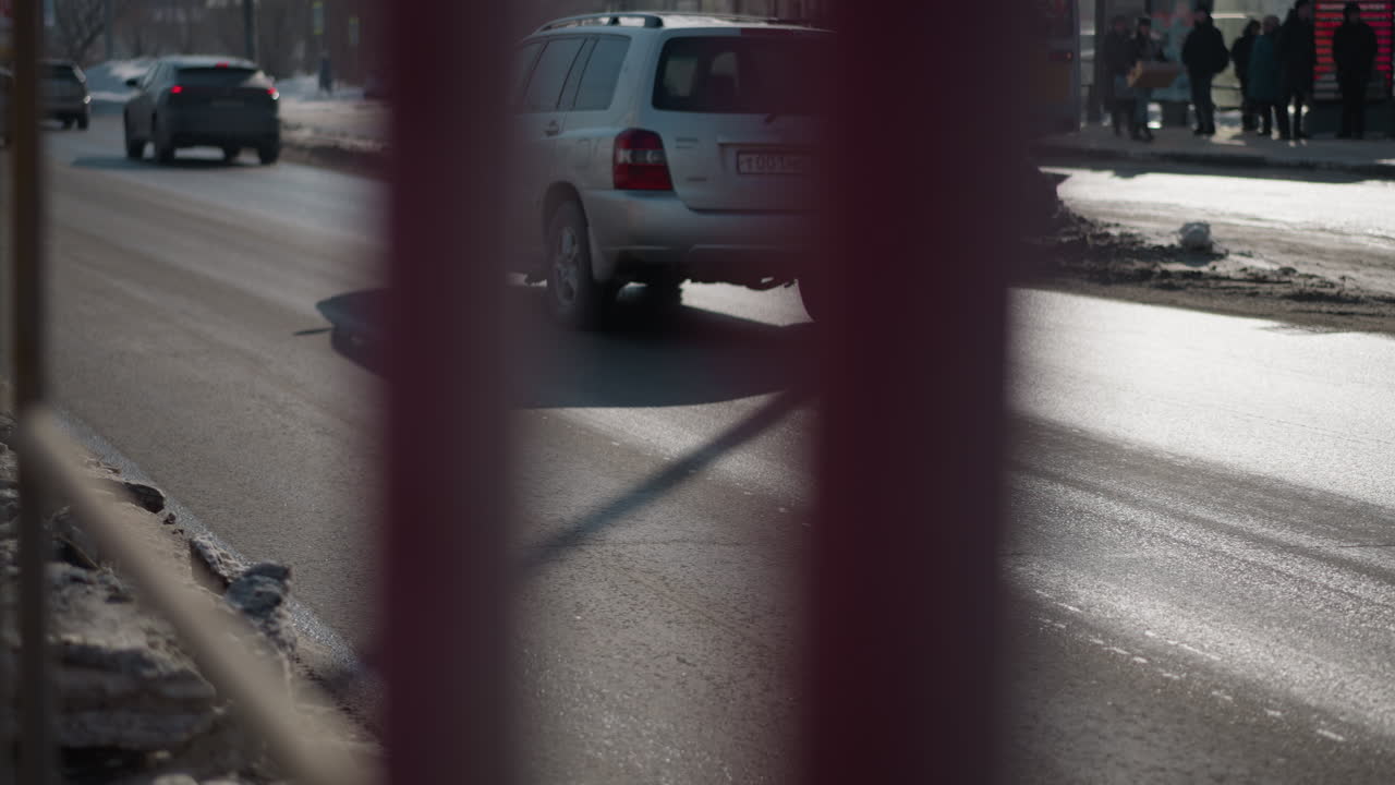 busy city road, cars moving along two lanes in winter sunlight, wet asphalt with glare, snow banks and parked vehicles in distance, side fence blur in foreground, urban commute scene, morning