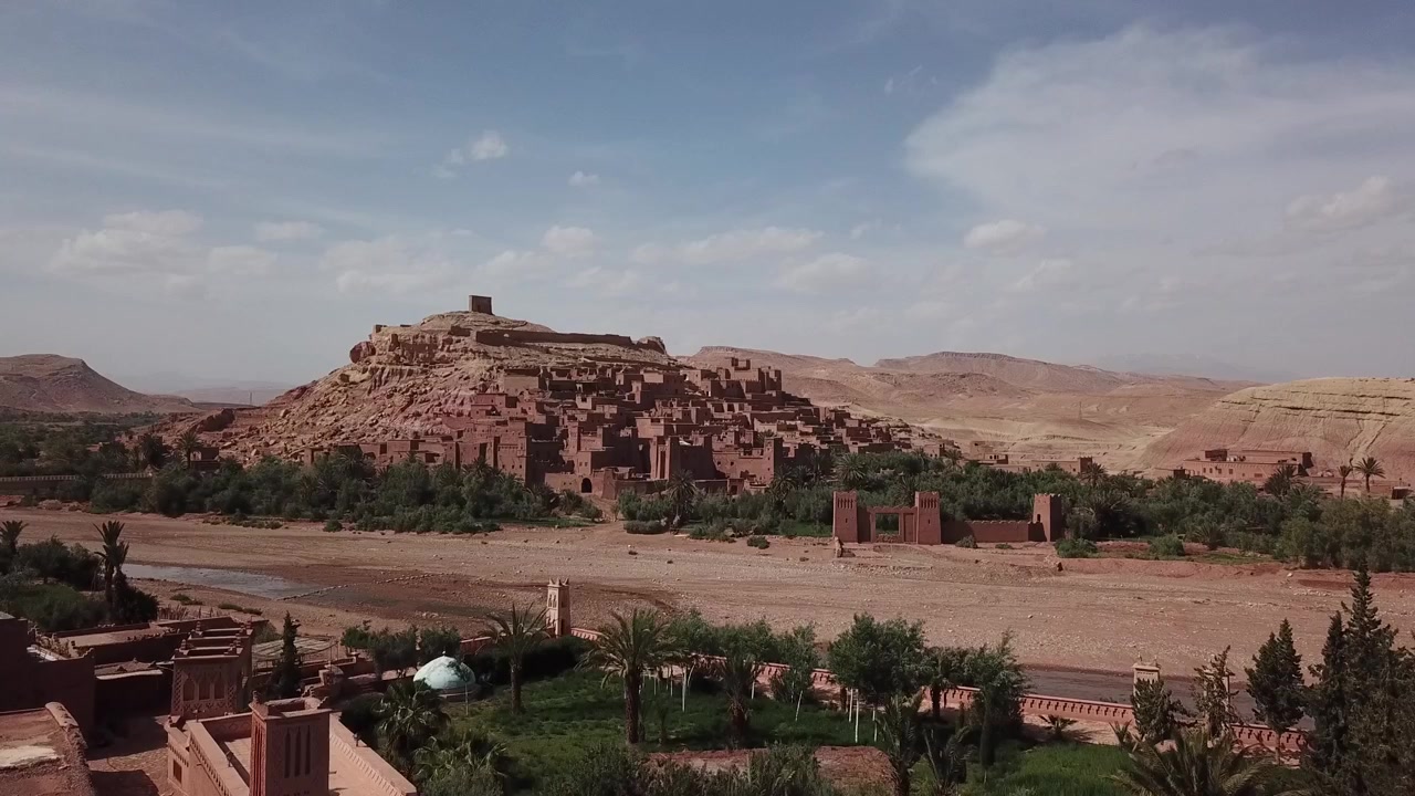 Naturskøn udsigt over palmer, der indrammer den historiske Ait Benhaddou kasbah i Ouarzazate, Marokko. Stærk forgrunds- og baggrundskomposition skaber en filmisk rejse- og kulturscene.