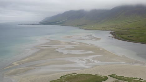 Aerial-approaching-shot-of-sandy-reef-of-Flateyri-with-green-coastline-during-cloudy-day,-Iceland