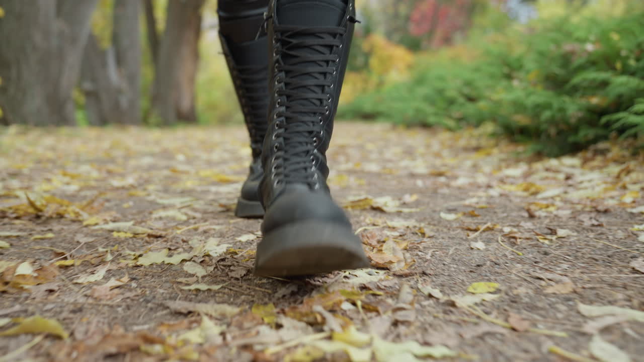 Nærbillede af sorte støvler, der spankulerer gennem parken, solbeskinnet sti, faldne blade, selvsikker pendlerstemning, fashionmodel-energi, lifestyleblogger-tempo, rytmiske skridt, kontrast mellem by og natur, afslappet