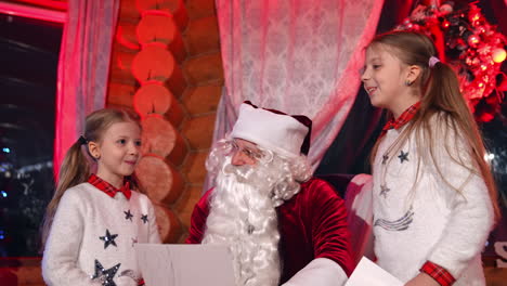 santa is resting in the armchair holding a letter and girls are beside him. santa is asking something from the letter. elder sister is smiling.