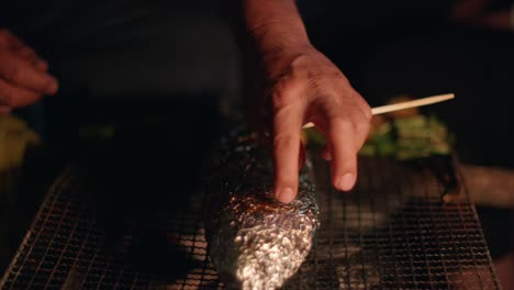 Pescado-Envuelto-En-Papel-De-Aluminio-Sentado-En-Una-Parrilla-Para-Cocinar-Siendo-Revisado-A-Mano-Para-Ver-Si-Está-Tierno,-Filmado-En-Cámara-Lenta