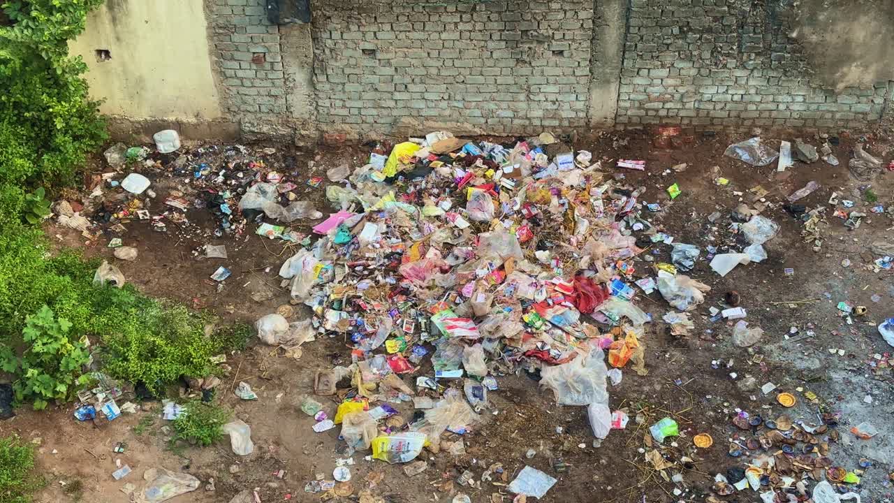 Aerial view of a pile of urban waste near a brick wall, showing mixed plastic, food, and paper trash — a stark reminder of poor waste disposal and rising environmental pollution