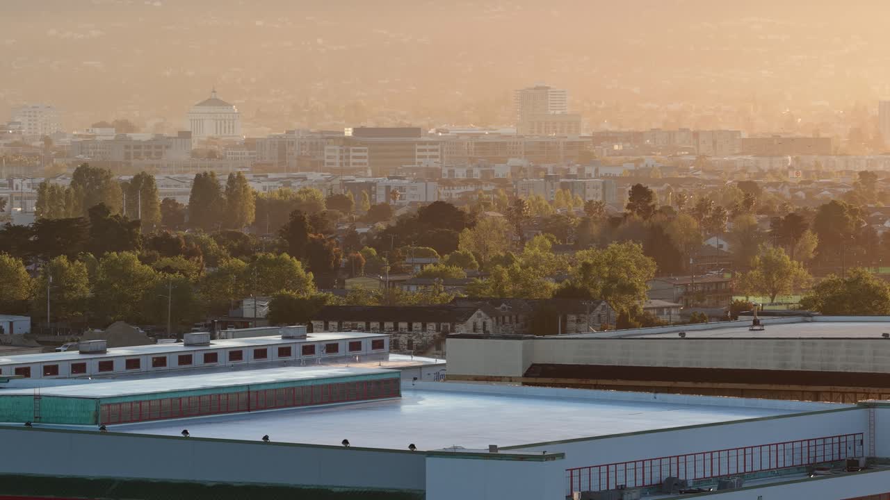 Aerial perspective revealing the city of Oakland through a warm, hazy sunset, with industrial rooftops in the foreground and the urban skyline emerging through the golden light